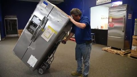 mover wheeling a stainless refrigerator strapped to an appliance hand truck, face visible