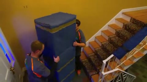two movers carrying a blanket-wrapped cabinet down a staircase, face visible