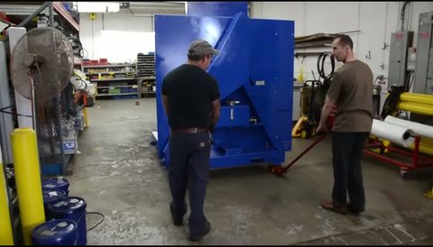 two-man crew rolling a large blue industrial machine across a shop floor on machinery skates with a steering bar, face visible