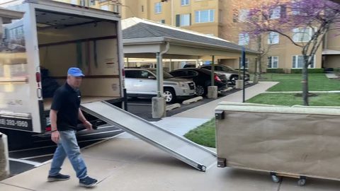 Wide shot of a mover walking past an open box truck at the curb with the loading ramp down and a wrapped dresser on a dolly, apartment...