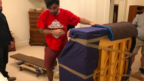 Mover strapping a blanket-wrapped upright piano onto a wheeled piano board while a second mover assists, face visible